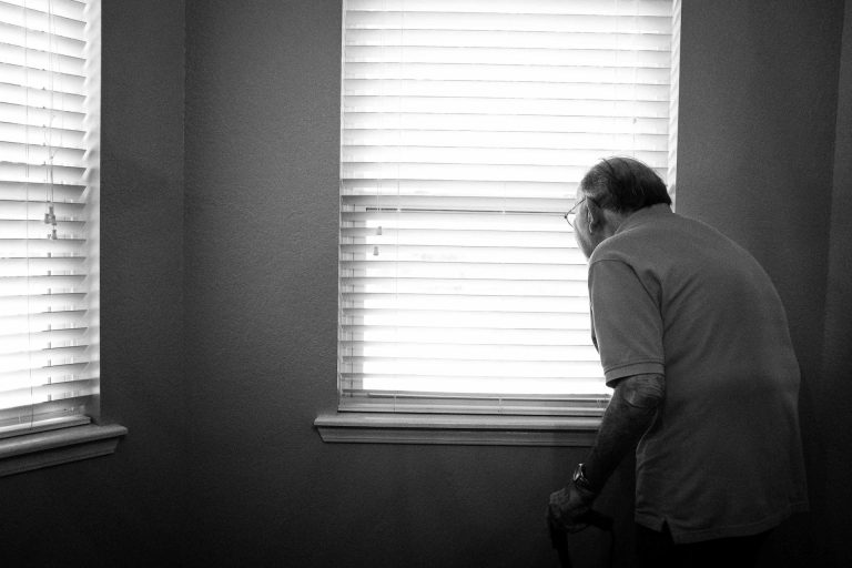 Photo en noir et blanc d’un homme âgé, appuyé sur une canne, qui regarde par des stores vénitiens entrebâillés. Lumière forte venant de la fenêtre, ambiance calme et un peu mélancolique.