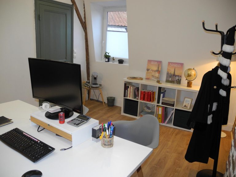 Bureau d’avocat cosy et lumineux : grand desk avec écran et clavier, pot à stylos, calculatrice; bibliothèque basse avec livres, globe et affiches “Arras” et “Bruxelles”; fenêtre mansardée, parquet clair, porte vert gris, et toge d’avocat accrochée sur un portemanteau.