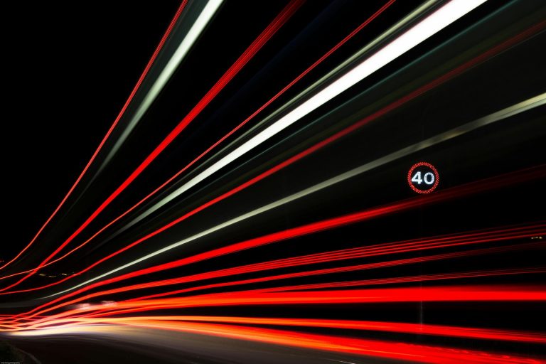 Longue exposition de traînées lumineuses rouges et blanches sur une route de nuit, avec un panneau de limitation à 40 km/h visible sur la droite.